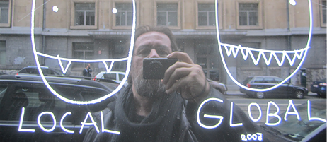 Dan Perjovschi macht in einem spiegelnden Fenster ein Selfie; auf dem Fenster sind links und rechts Köpfe skizziert mit den Unterschriften local und global