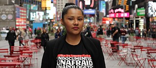 A woman stands in Time Square, wearing a t-shirt that says "undocumented, unafraid, unapologetic."