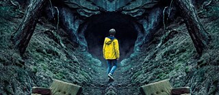 A young man wearing a yellow raincoat at the entrance of a cave in a forest