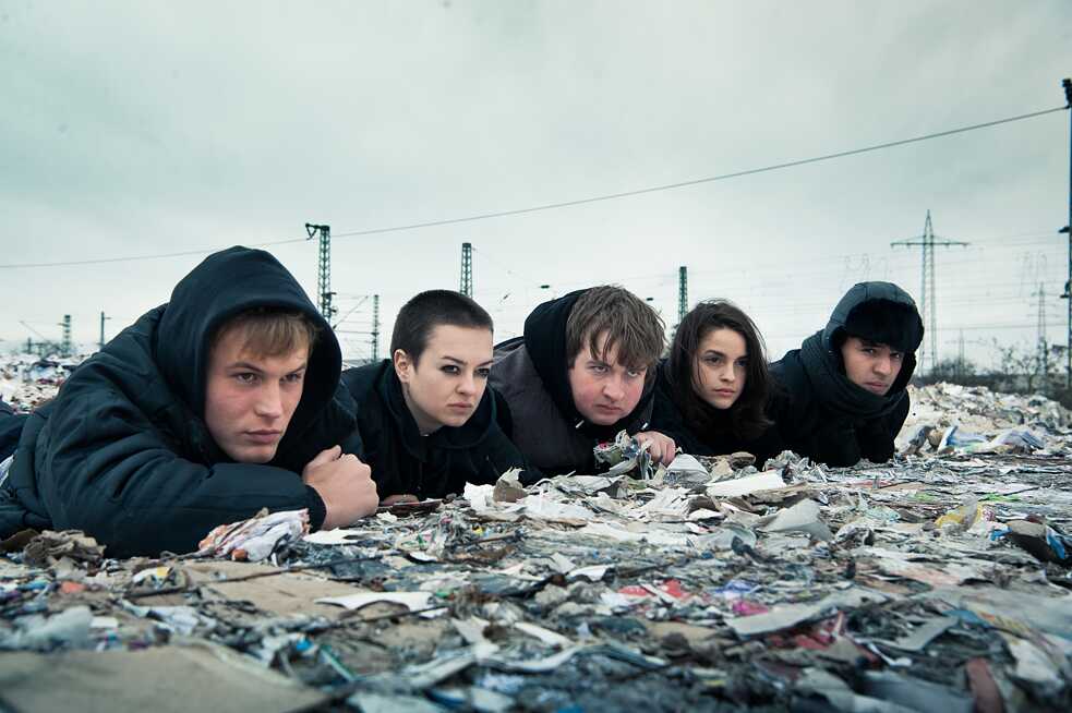 Standbild aus der Netflix Deutschland Serie "We are the wave/ Wir sind die Welle" Die Guppe der fünf jugendlichen Protagonisten lauert versteckt auf Halden von Altpapierballen.