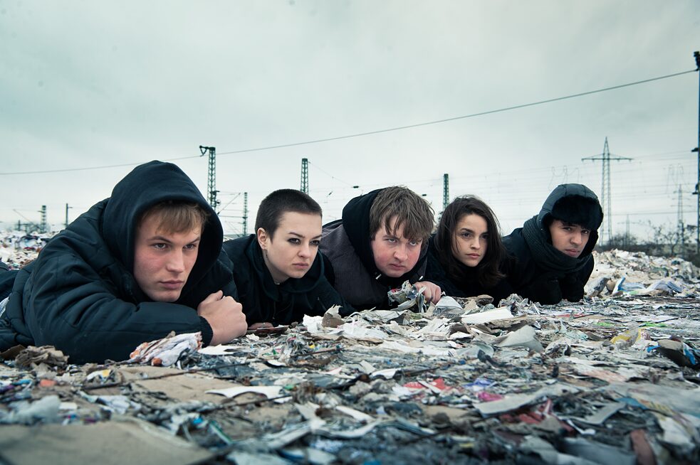 Still frame from the Netflix Germany original series "We are the wave": The group of five teenagers lying in wait on top of large waste paper bails.